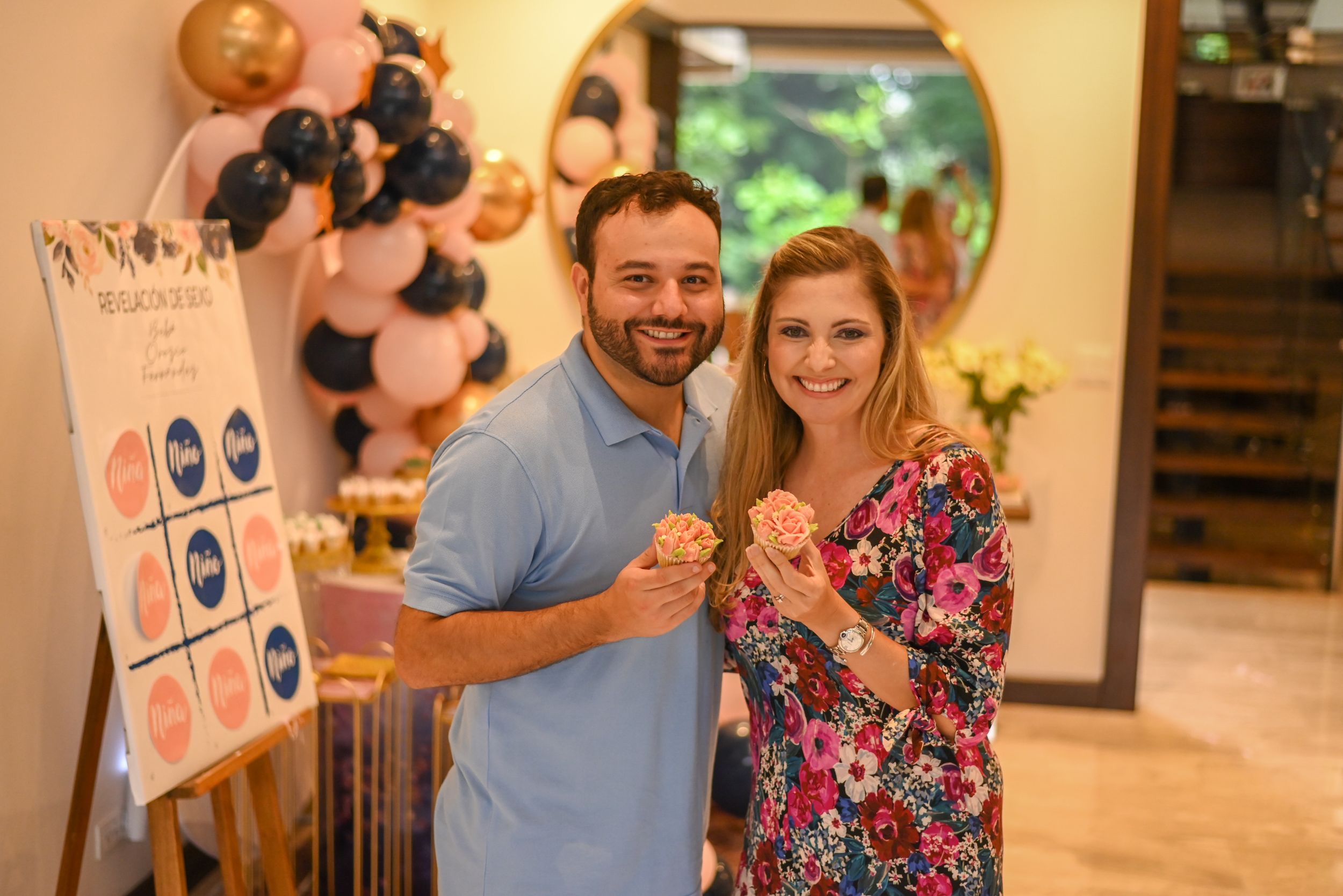 Couple smiles while holding donuts at a festive event with balloons and decorations in the background.