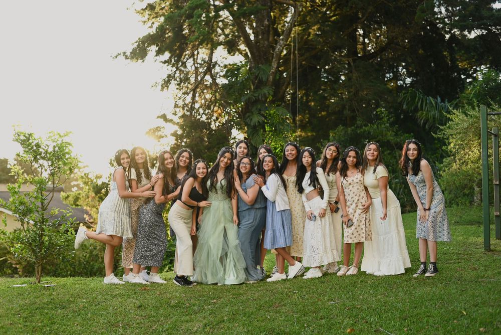 A group in light-colored dresses stands together on a grassy lawn surrounded by trees during a summer celebration.