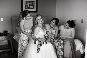 A black and white photo shows bridal party members in floral dresses sharing a laughing moment before the ceremony.