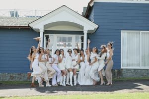 A large group wearing all white outfits poses together on the front porch steps of a blue house.