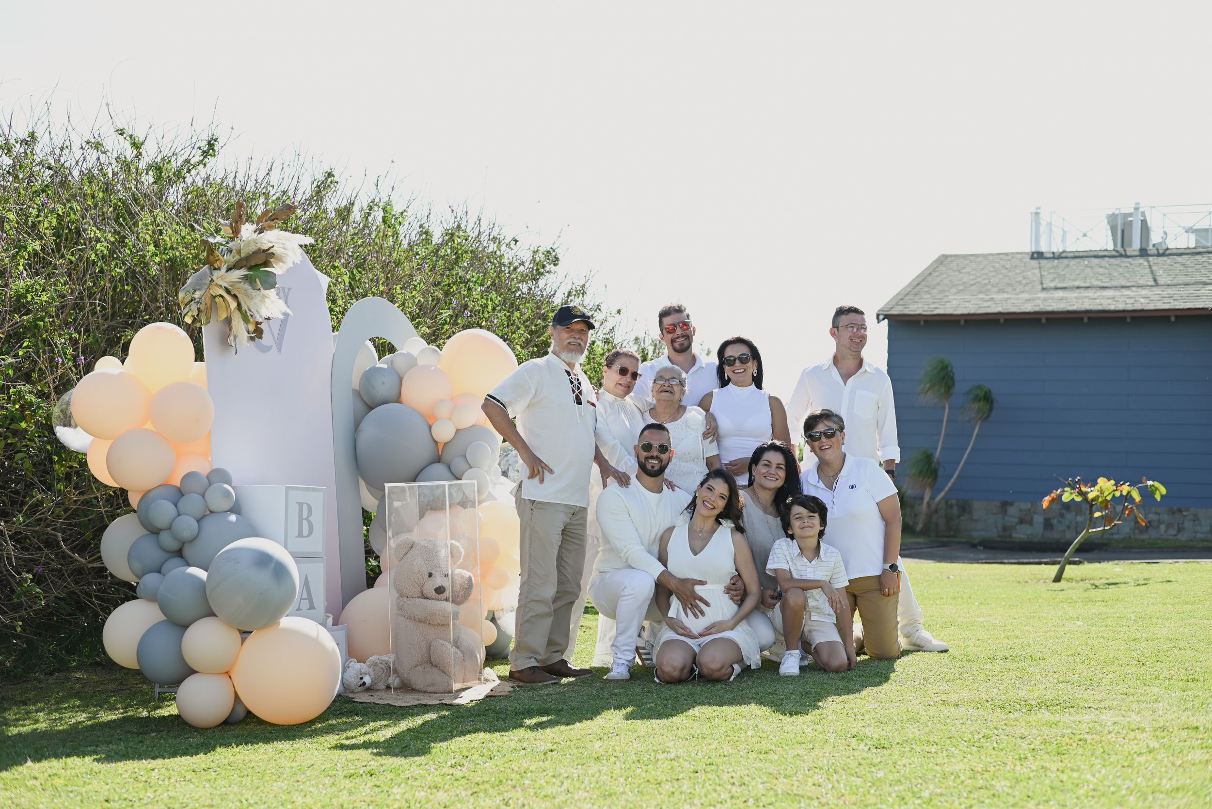 Outdoor celebration setup with white and peach balloon arch, decorative teddy bears, and group gathered in all-white attire on lawn.