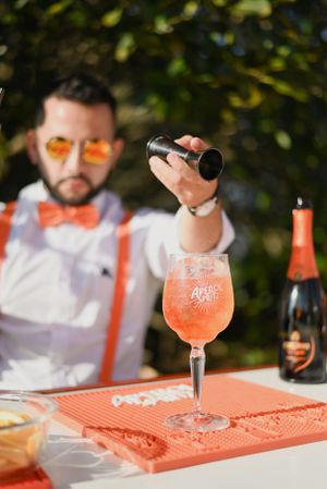 A bartender prepares a pink cocktail with bitters on an orange bar mat while holding a jigger to measure ingredients.