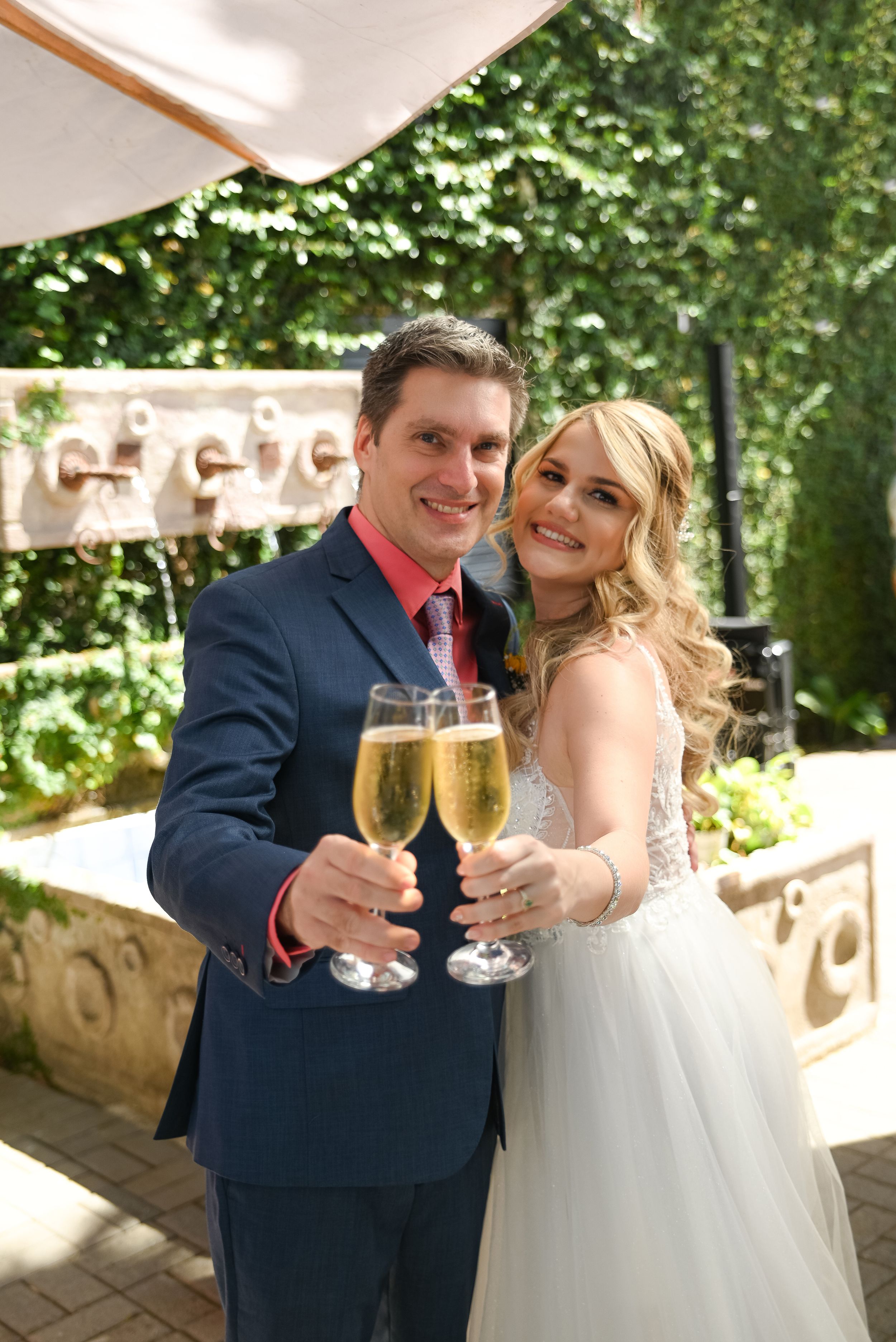 A bride and groom toast with champagne flutes at an outdoor wedding reception surrounded by greenery.