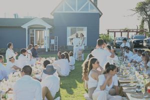 Wedding guests seated at long tables during an outdoor reception by a blue house on a sunny day.