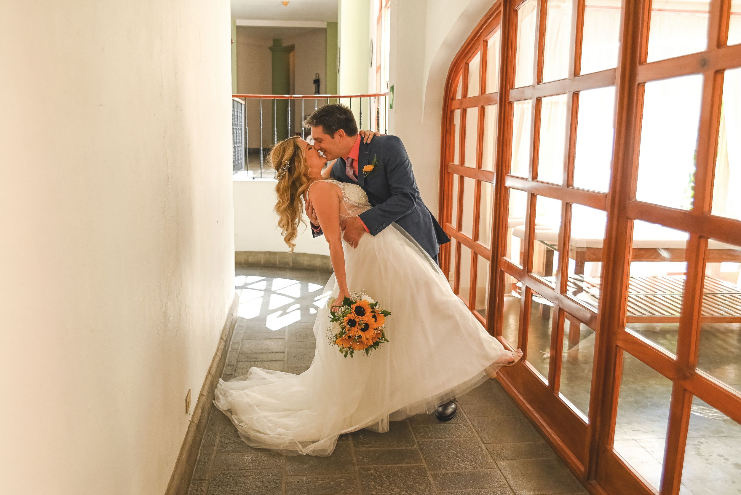 A couple dips romantically in front of sunlit arched windows with fall flowers in a white wedding dress and suit.