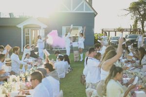 Guests enjoy an elegant outdoor dinner party on a lawn at sunset with white decor and string lights.