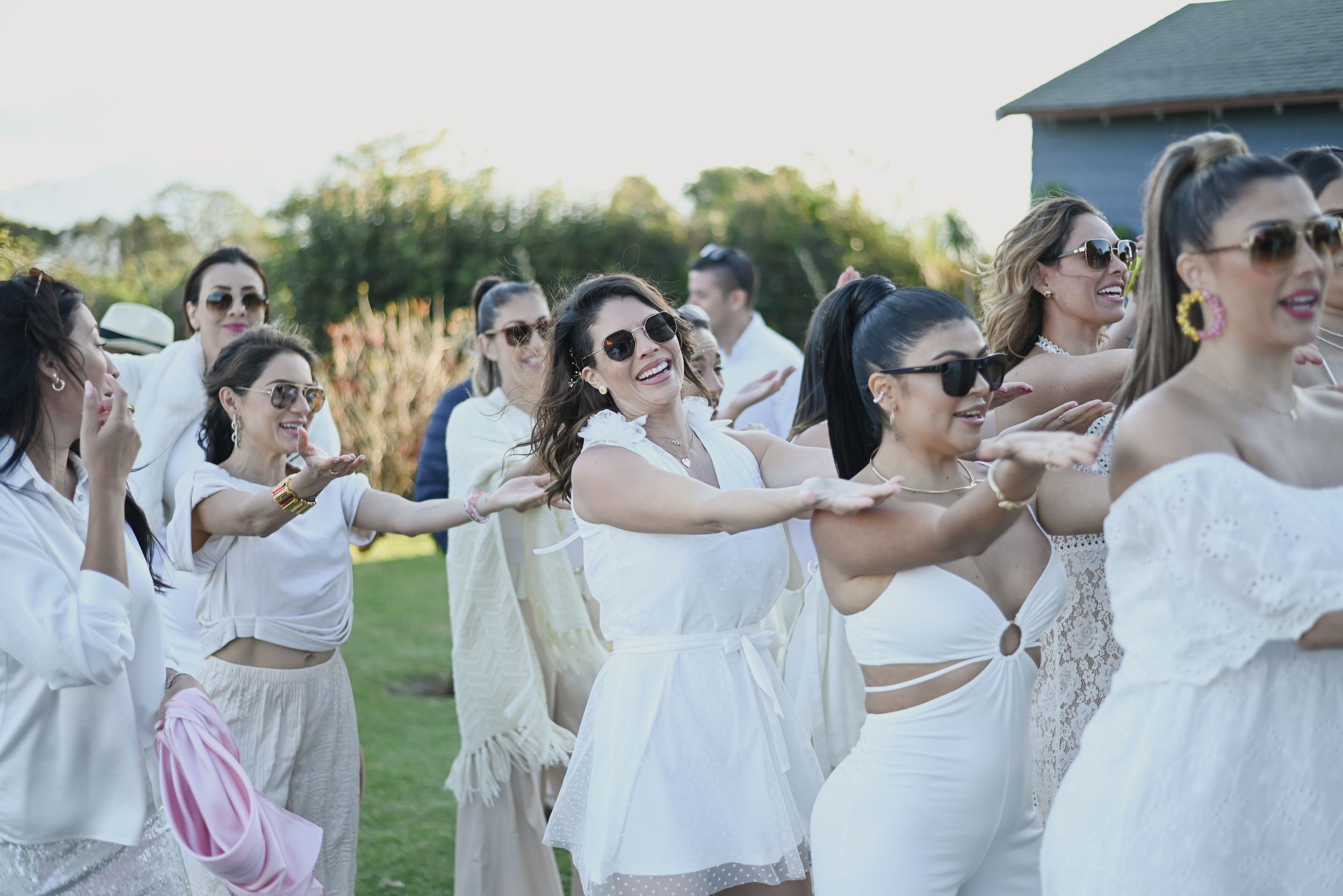 A group in white dresses and sunglasses dances together at an outdoor party during a sunny afternoon.