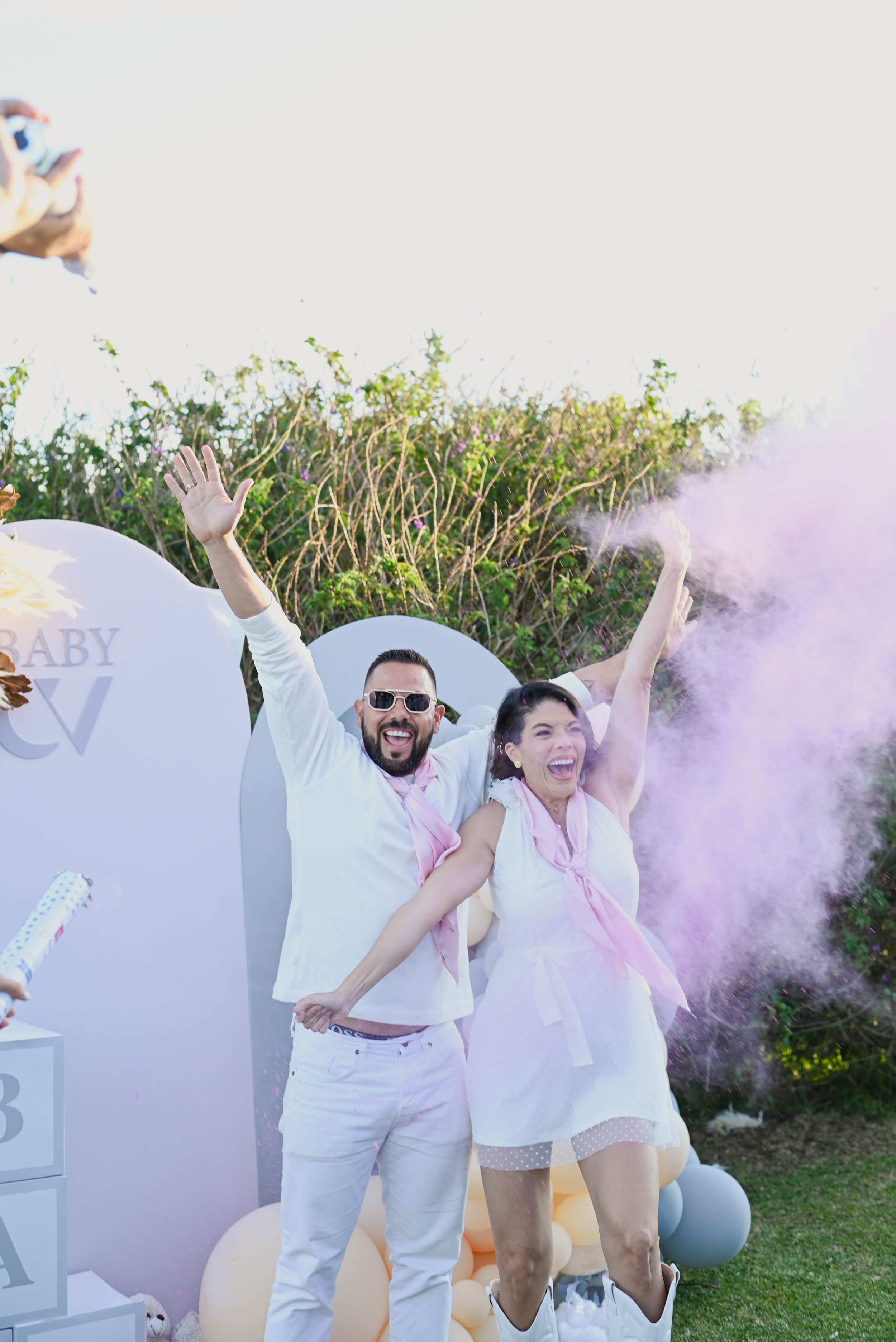 Couple celebrates with joy amidst pink smoke and baby shower decorations in an outdoor garden setting.