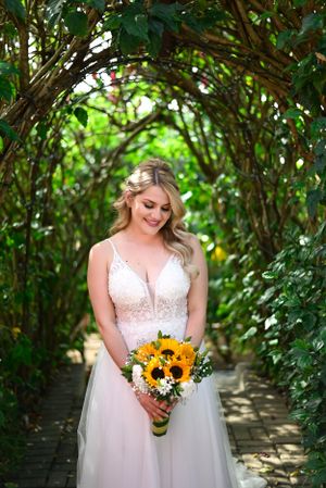 A bride in a white lace wedding gown holds a vibrant sunflower bouquet while standing under a garden archway.