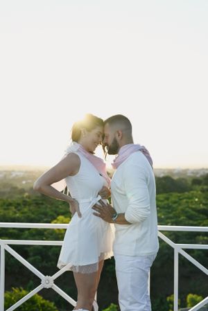 A romantic couple in white outfits embrace on a balcony at sunset with greenery and scenic views in the background.