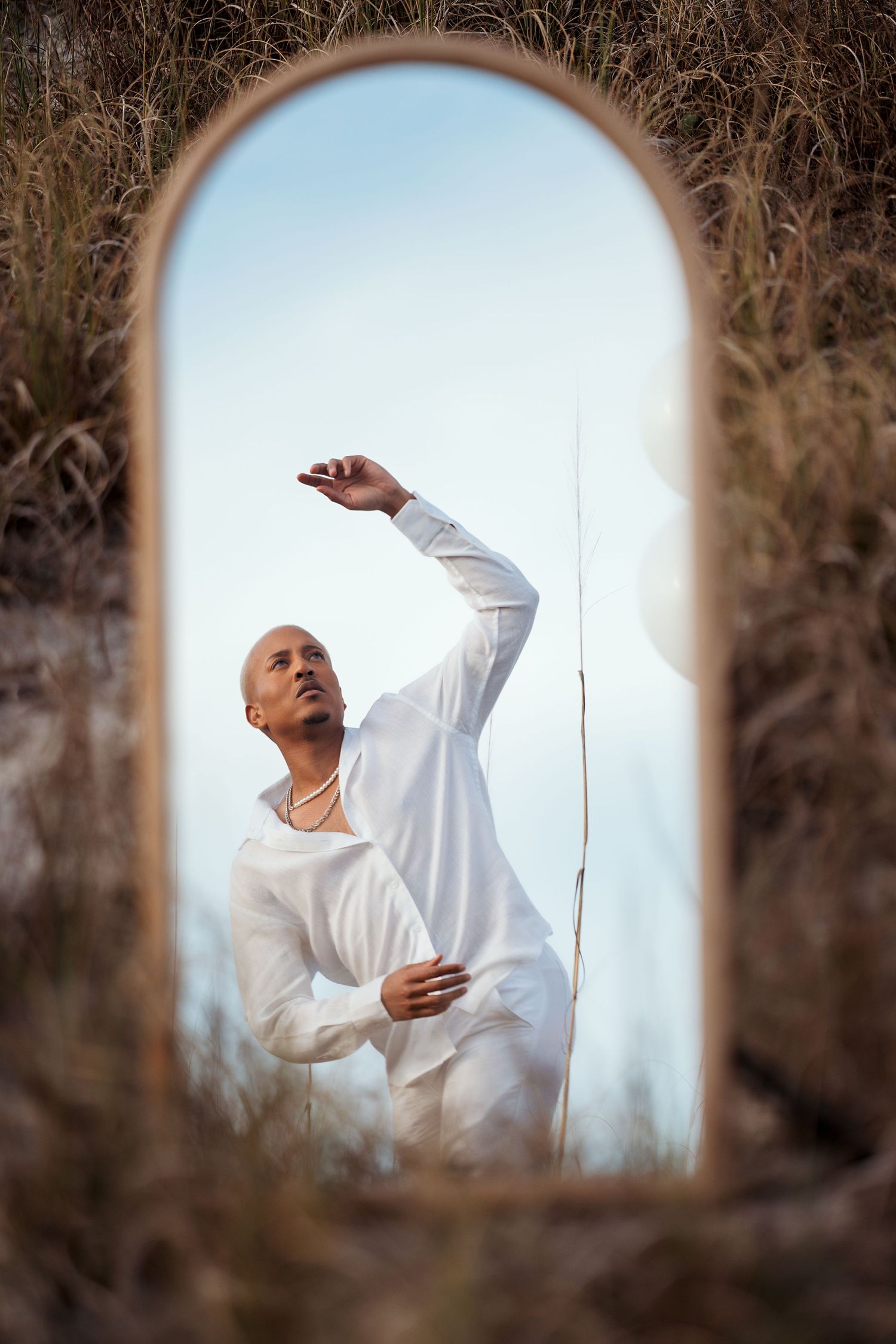 Artistic portrait through a round mirror frame shows a person in white clothing making an elegant dance pose in nature.