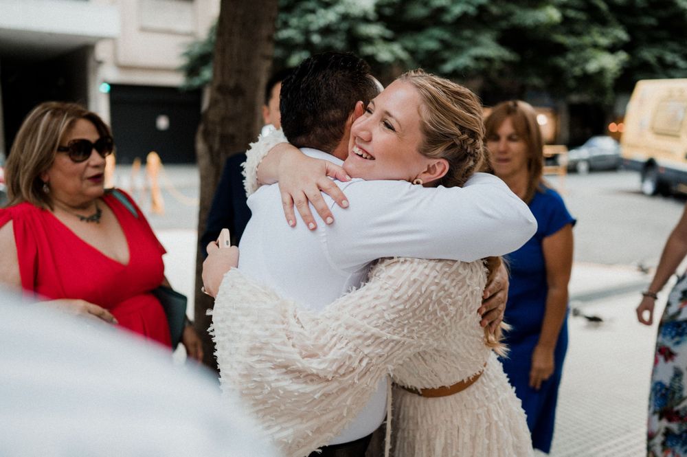 Two people share an emotional embrace while wearing wedding attire as others look on at an outdoor gathering.