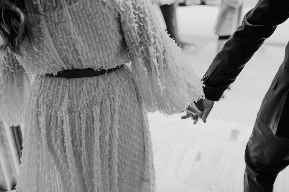 A close-up detail shot of hands reaching for each other, with a white dress visible in the frame.