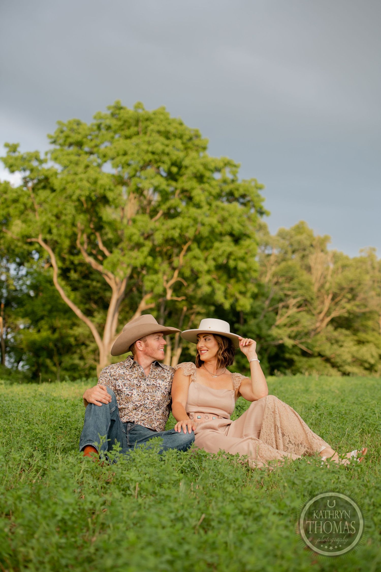 A couple in cowboy hats sitting on a hill with a dramatic cloudy sky behind them.