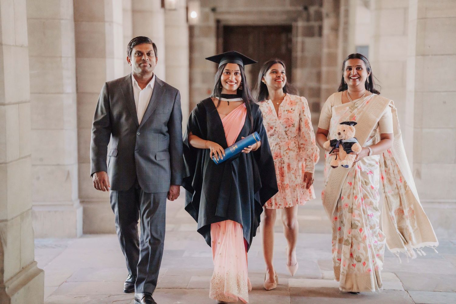 Group walks through elegant stone hallway in formal attire and graduation robes.