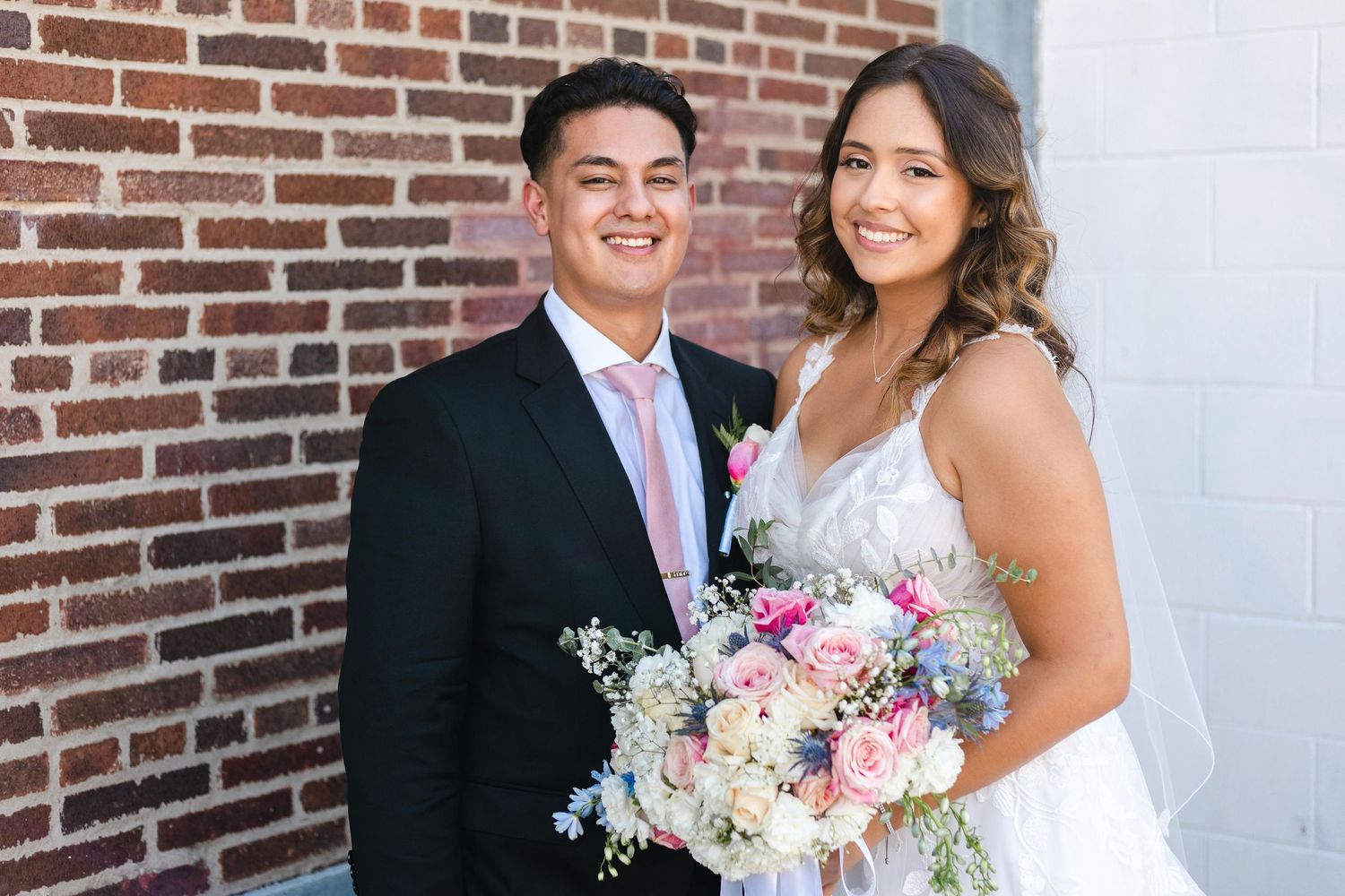 A newlywed couple smiles together against a brick wall while holding a beautiful pastel pink and white wedding bouquet.
