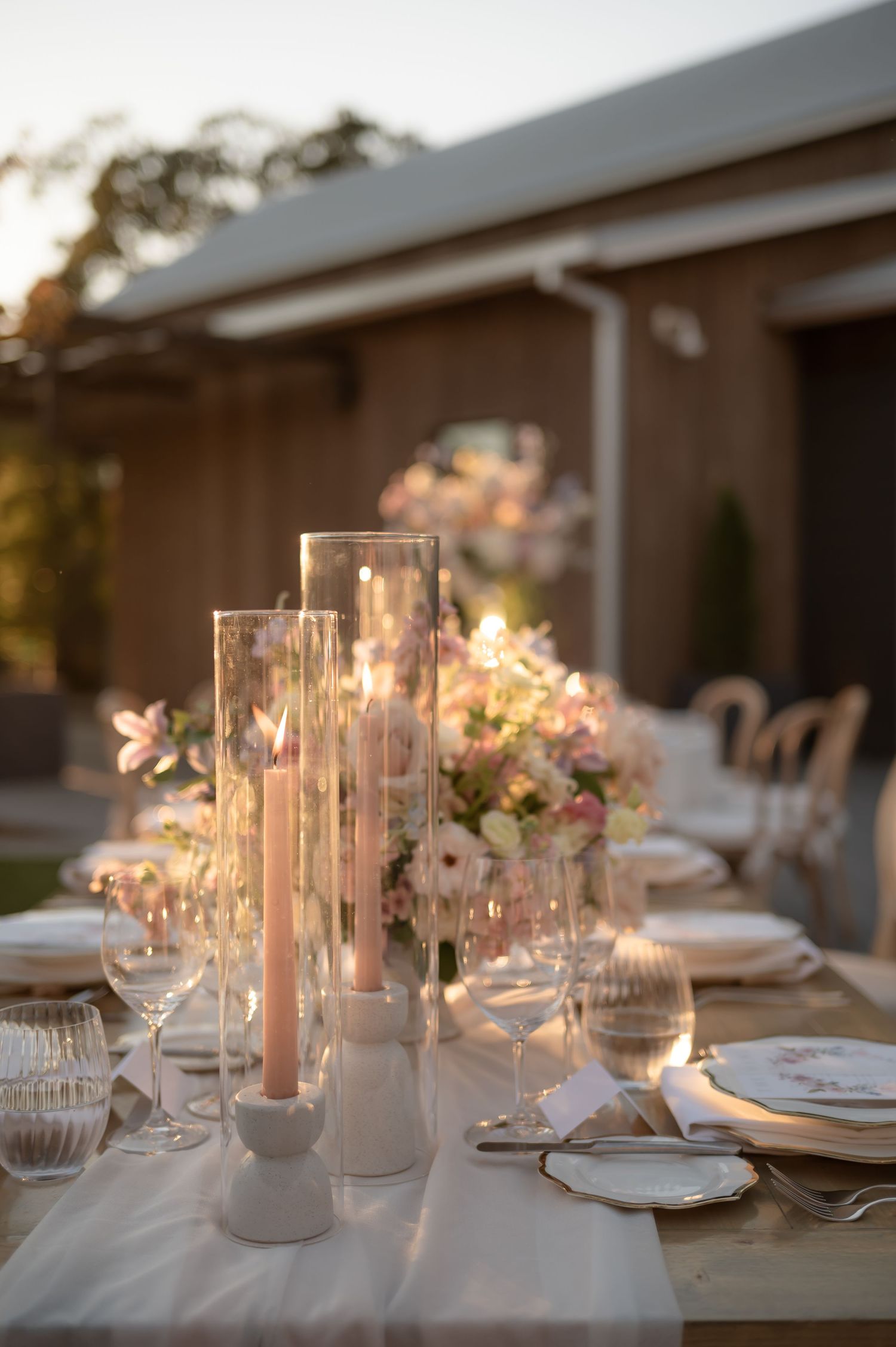 Elegant outdoor table setting with tall glass candle holders and pink floral arrangements glowing in sunset light.