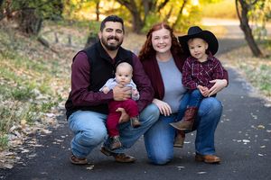 A family of four poses together for family photographer outdoors during autumn with fallen leaves and golden sunlight in background.
