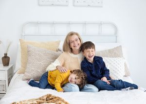 Three people sit together and smile on a white bed with textured pillows and blankets in a bright room.