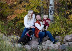 Three people sit together on rocks surrounded by autumn foliage and birch trees in a natural outdoor setting.