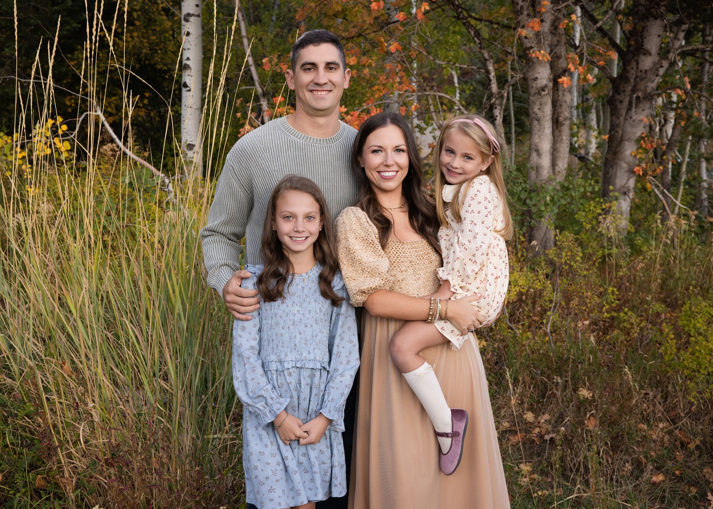 A family stands together in an outdoor setting with tall grass and autumn foliage in the background.