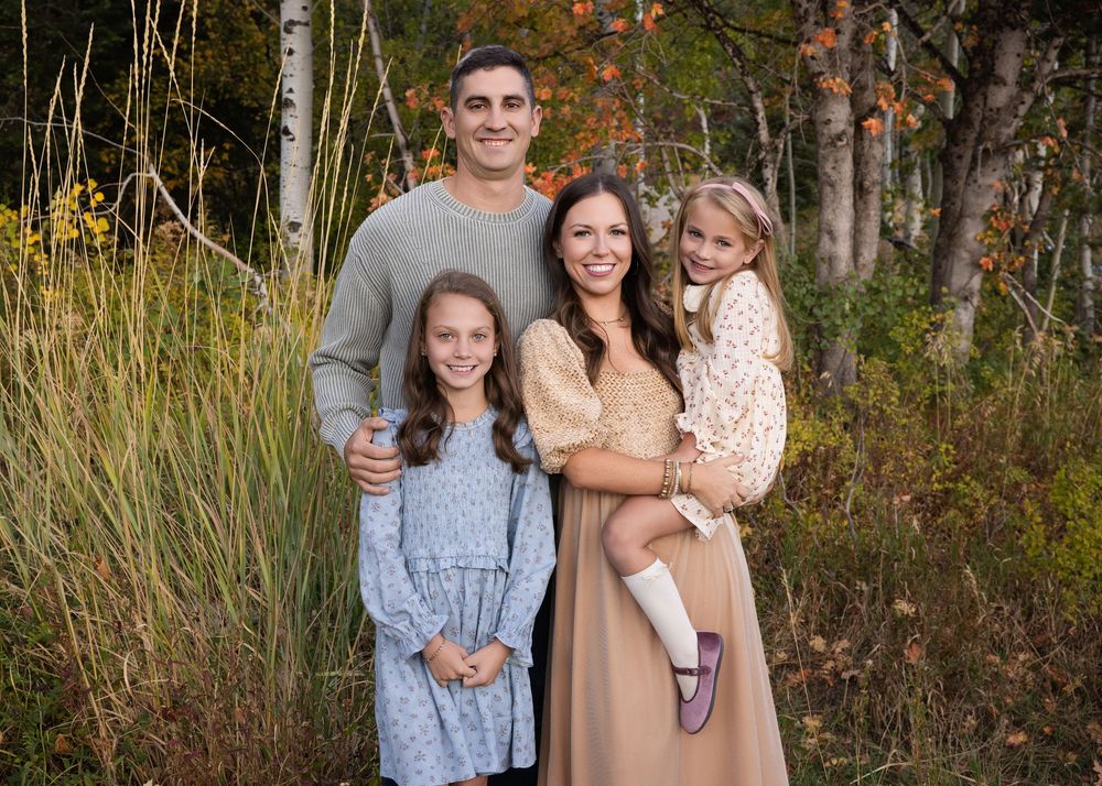 A family stands together in an outdoor setting with tall grass and autumn foliage in the background.