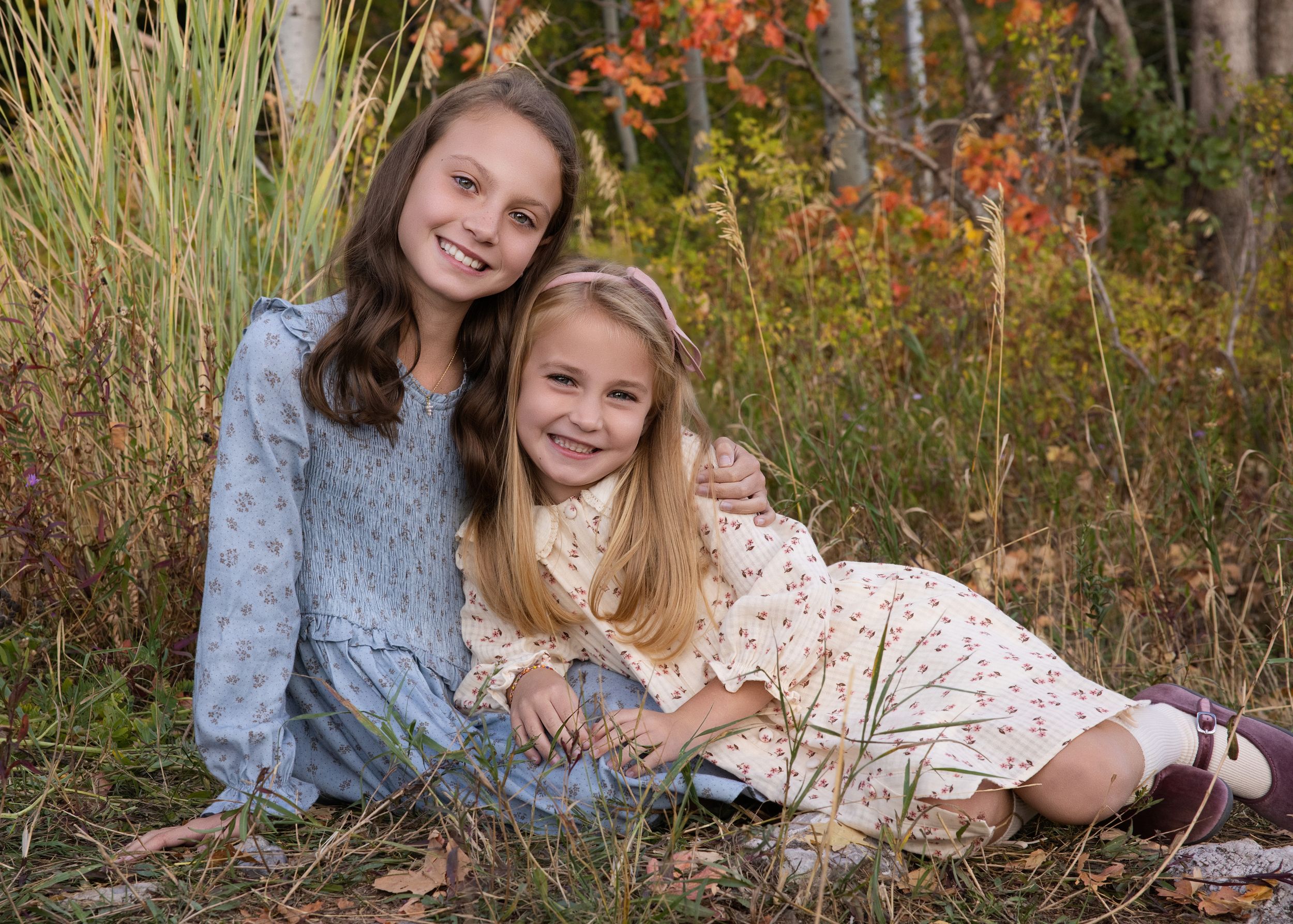 Two children in dresses sitting together in tall autumn grass with colorful fall foliage in the background.