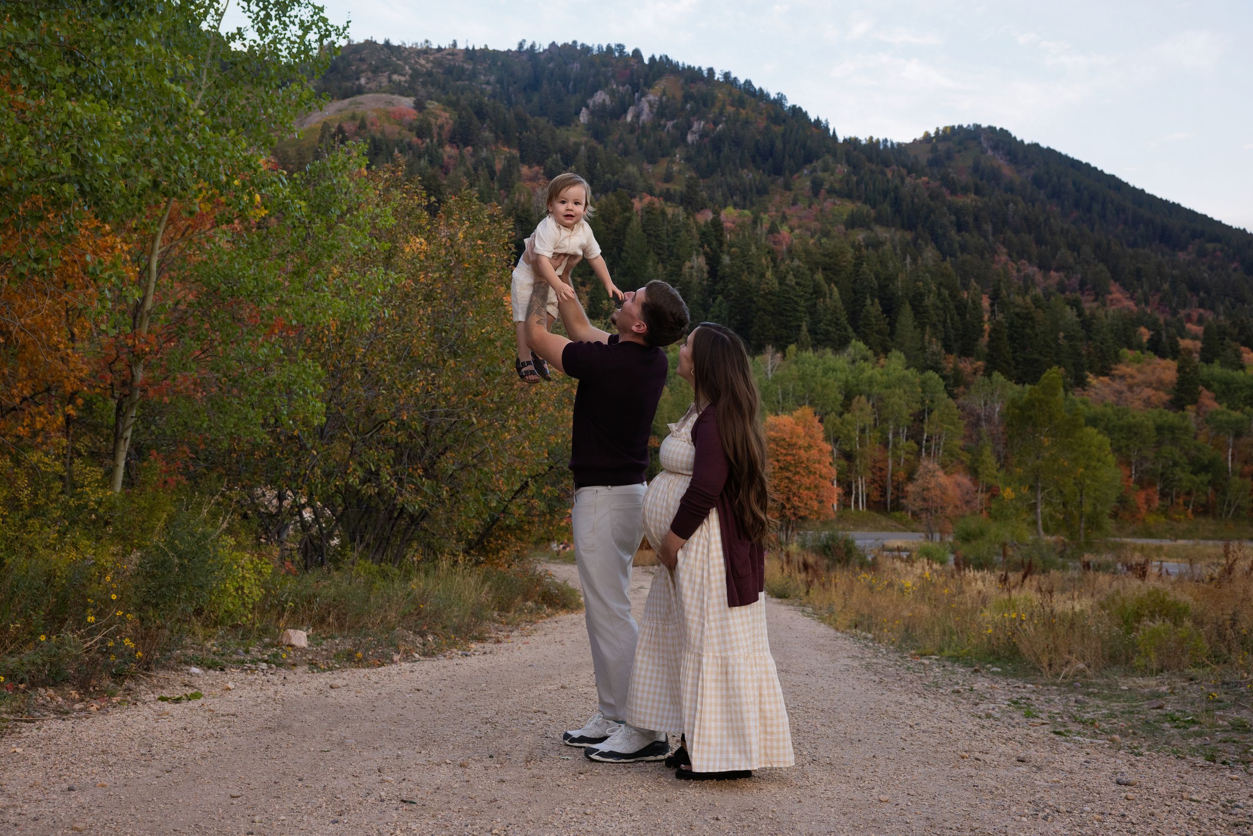Two people in formal attire walk along a mountain trail during autumn with colorful foliage in the background.