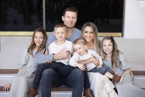 A family with three children sits together on white steps wearing coordinated neutral colored outfits.