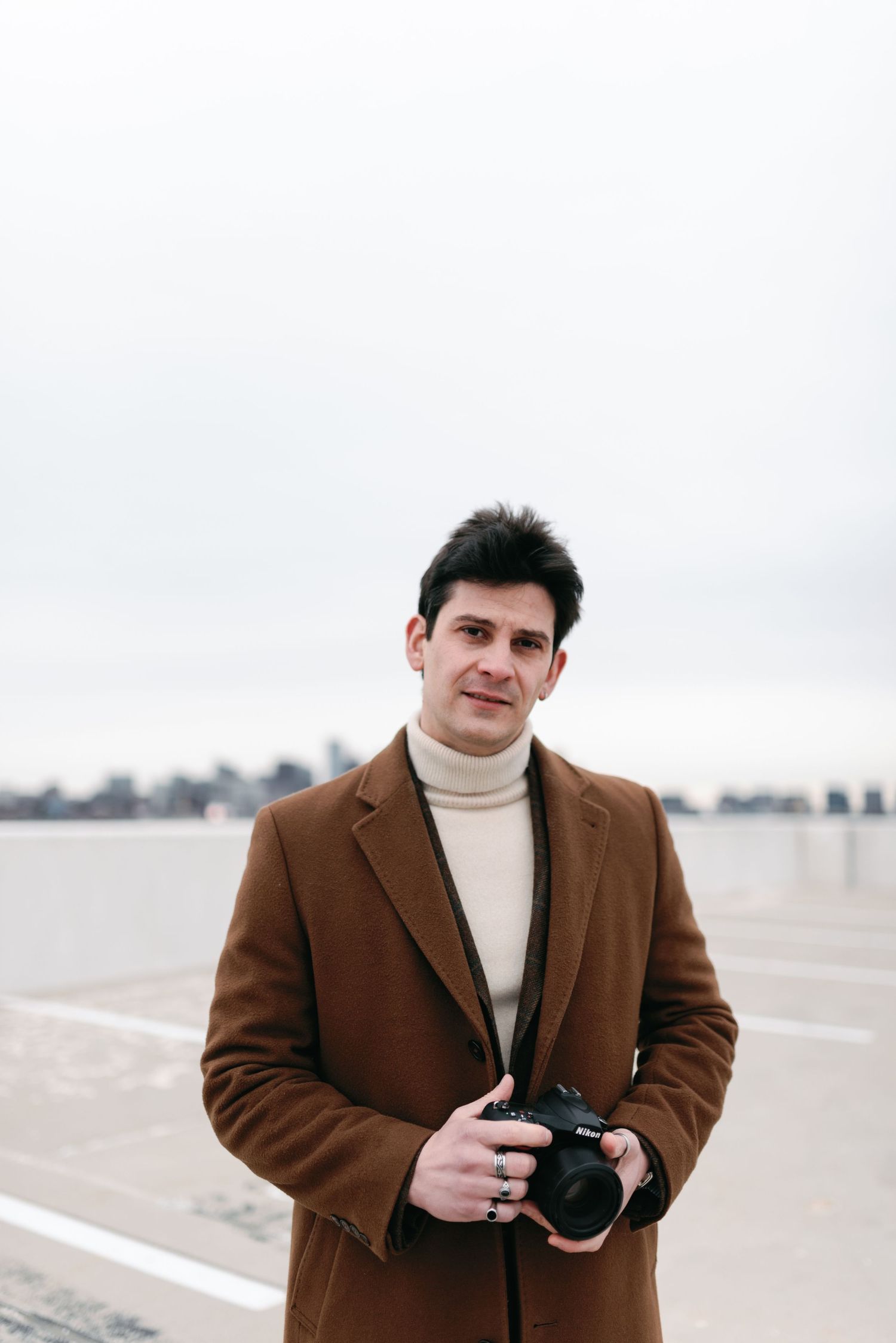 A portrait of a photographer in a turtleneck and brown coat holding a camera on an urban rooftop.