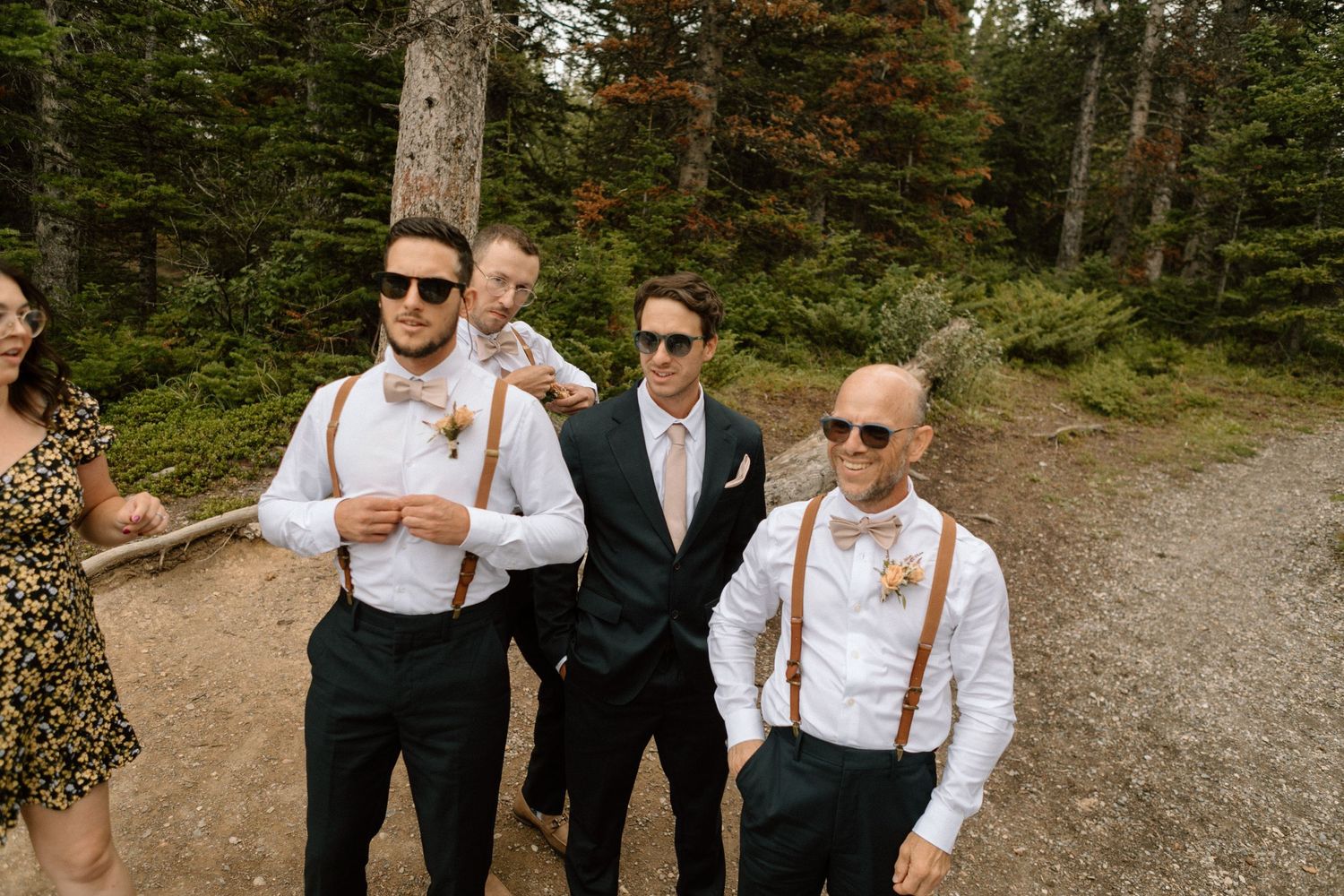Groomsmen in suspenders and sunglasses stand together on a mountain trail surrounded by evergreen forest.