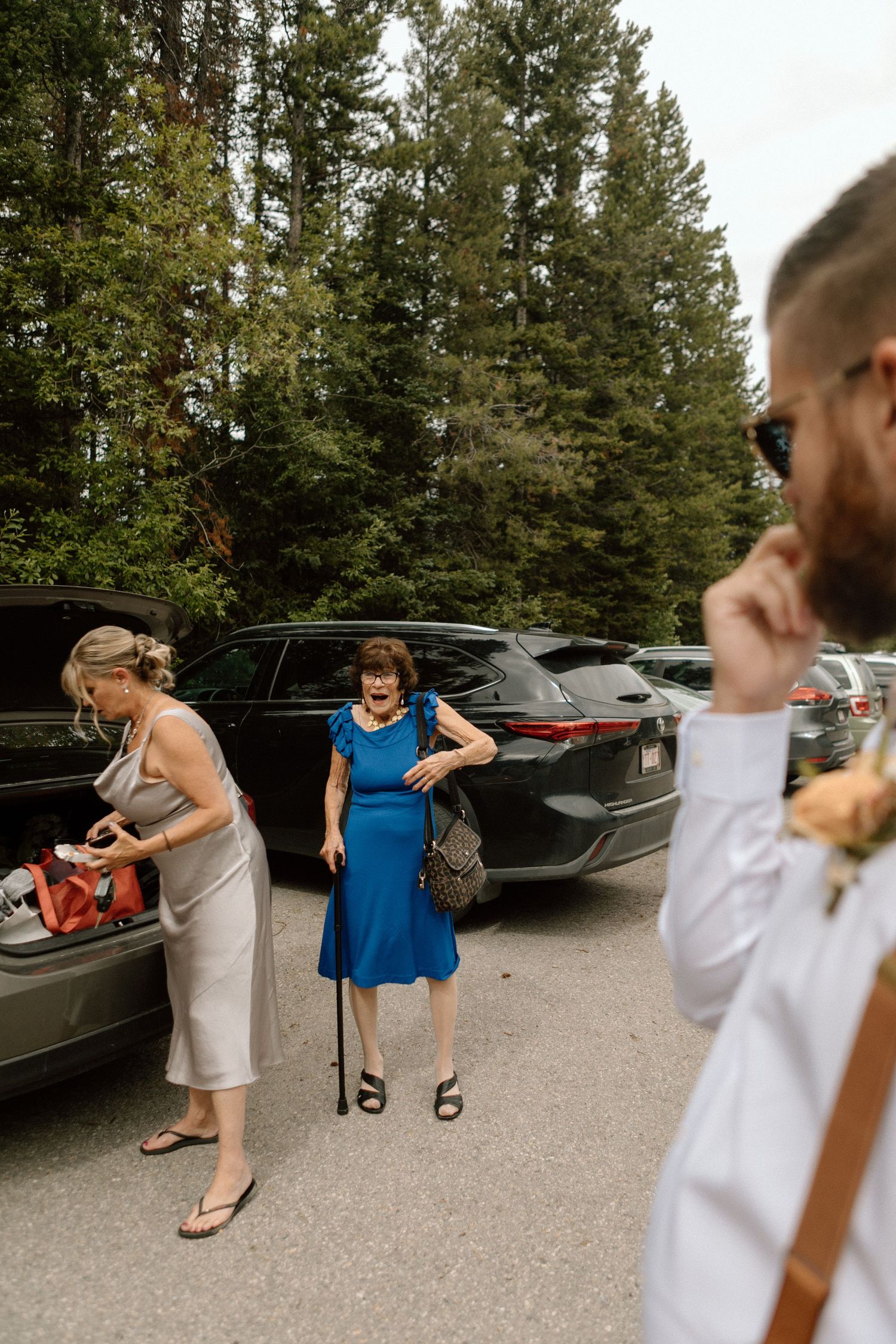 Wedding guests arrive and unload cars in a forest parking area on overcast day.