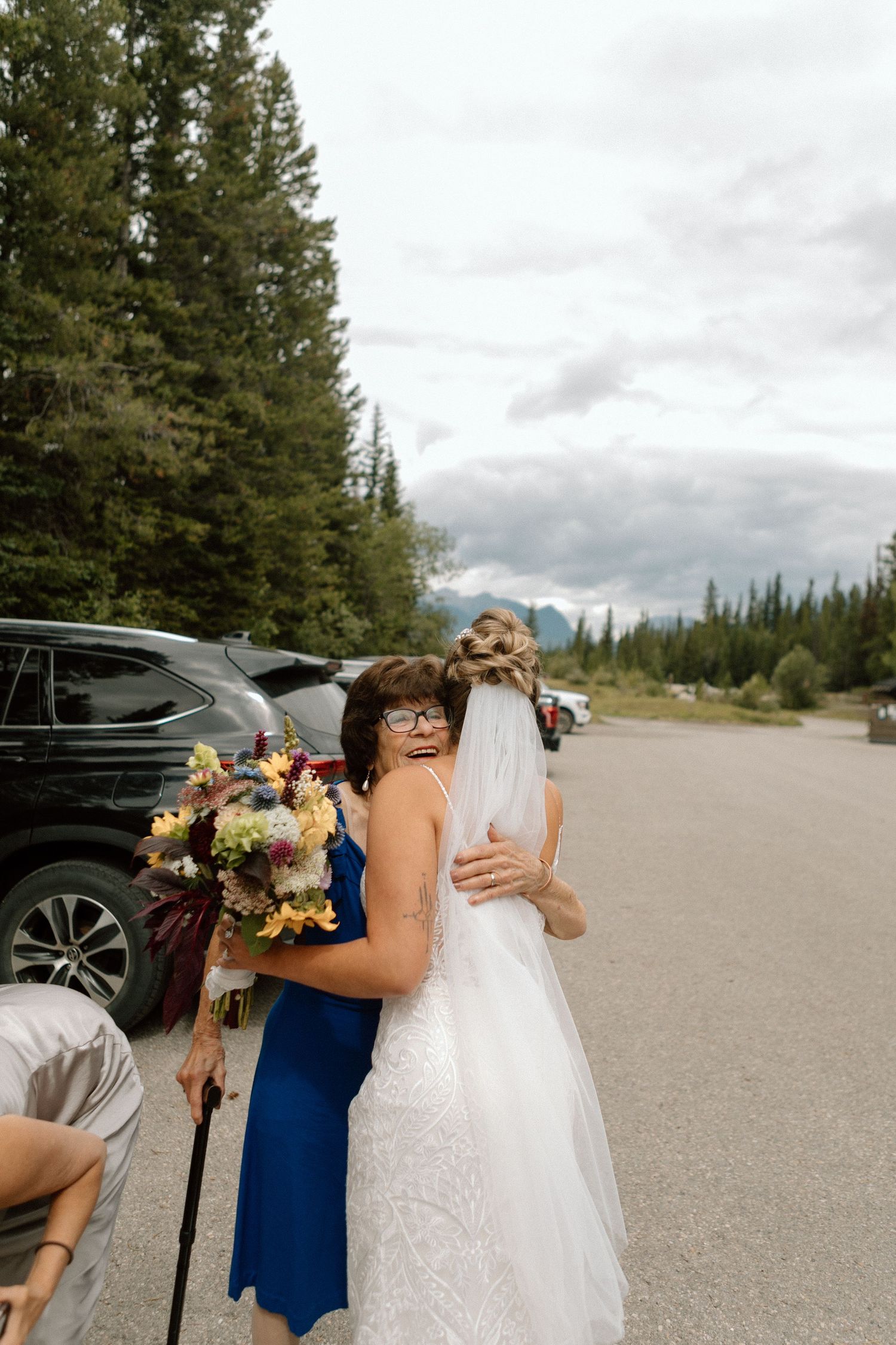 Emotional embrace between wedding guests in a mountainside parking lot with evergreen trees.