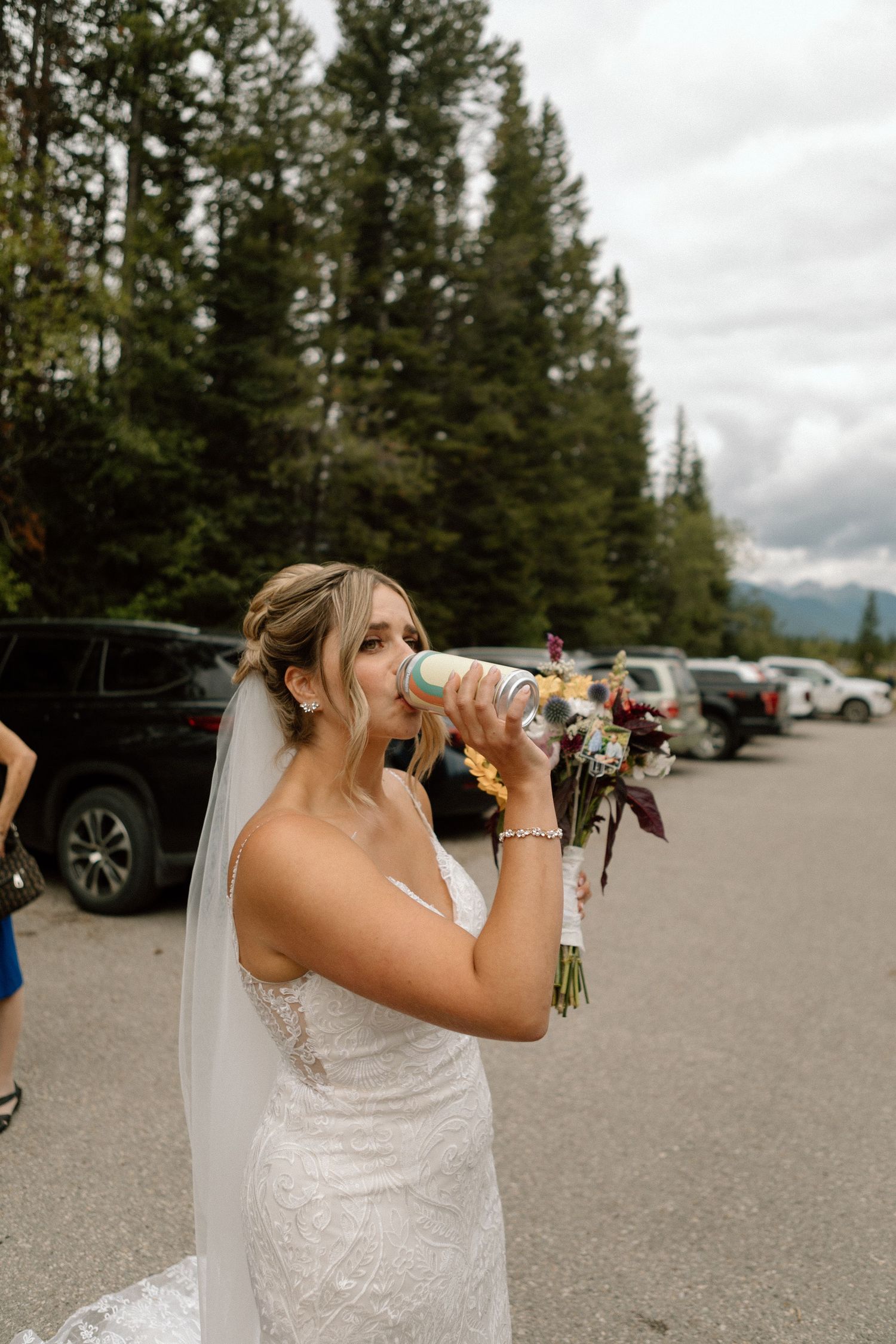 Bride in lace gown drinks from water bottle while holding bouquet in forest parking lot.