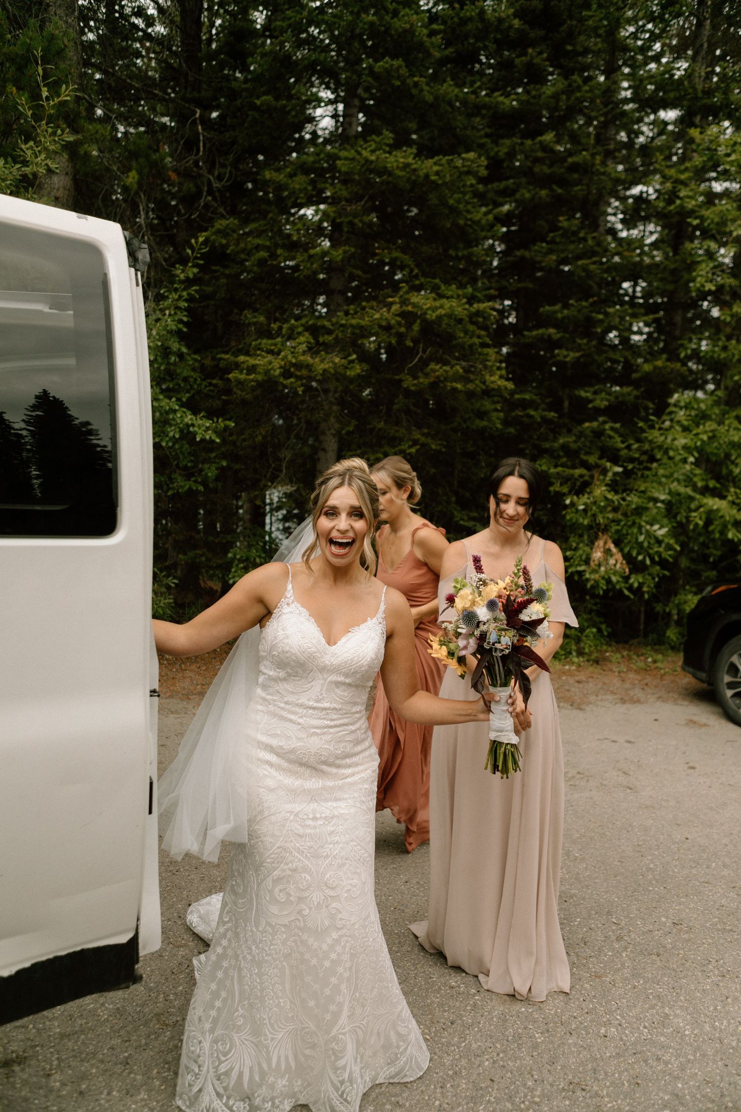 Wedding party exits white van on forest path with bride in lace dress and bridesmaids in blush gowns.