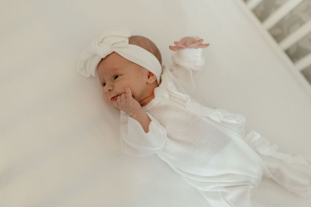 Baby in white outfit smiles while lying in crib. Virginia/ North Carolina Newborn In-Home Session