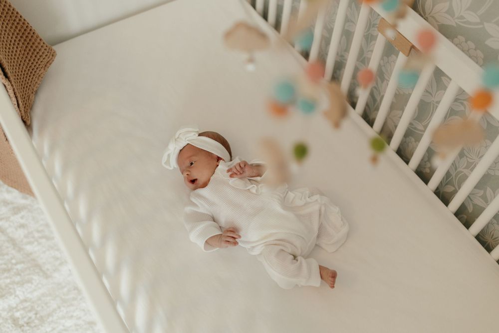 Baby in white clothing lies in crib under hanging mobile toys. Virginia/ North Carolina Newborn In-Home Session