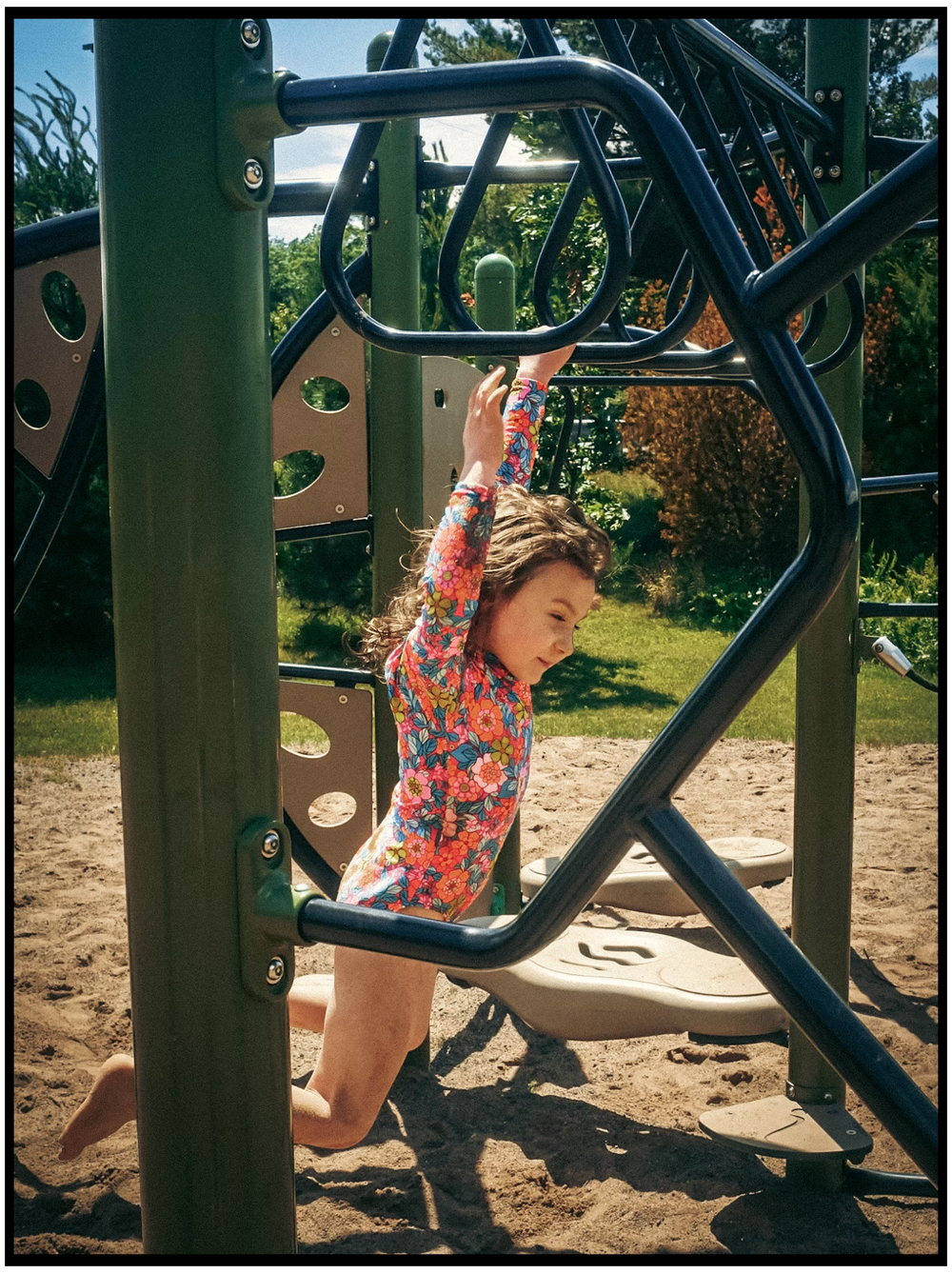 A child in a floral shirt plays on a modern playground climbing structure on a sunny day.
