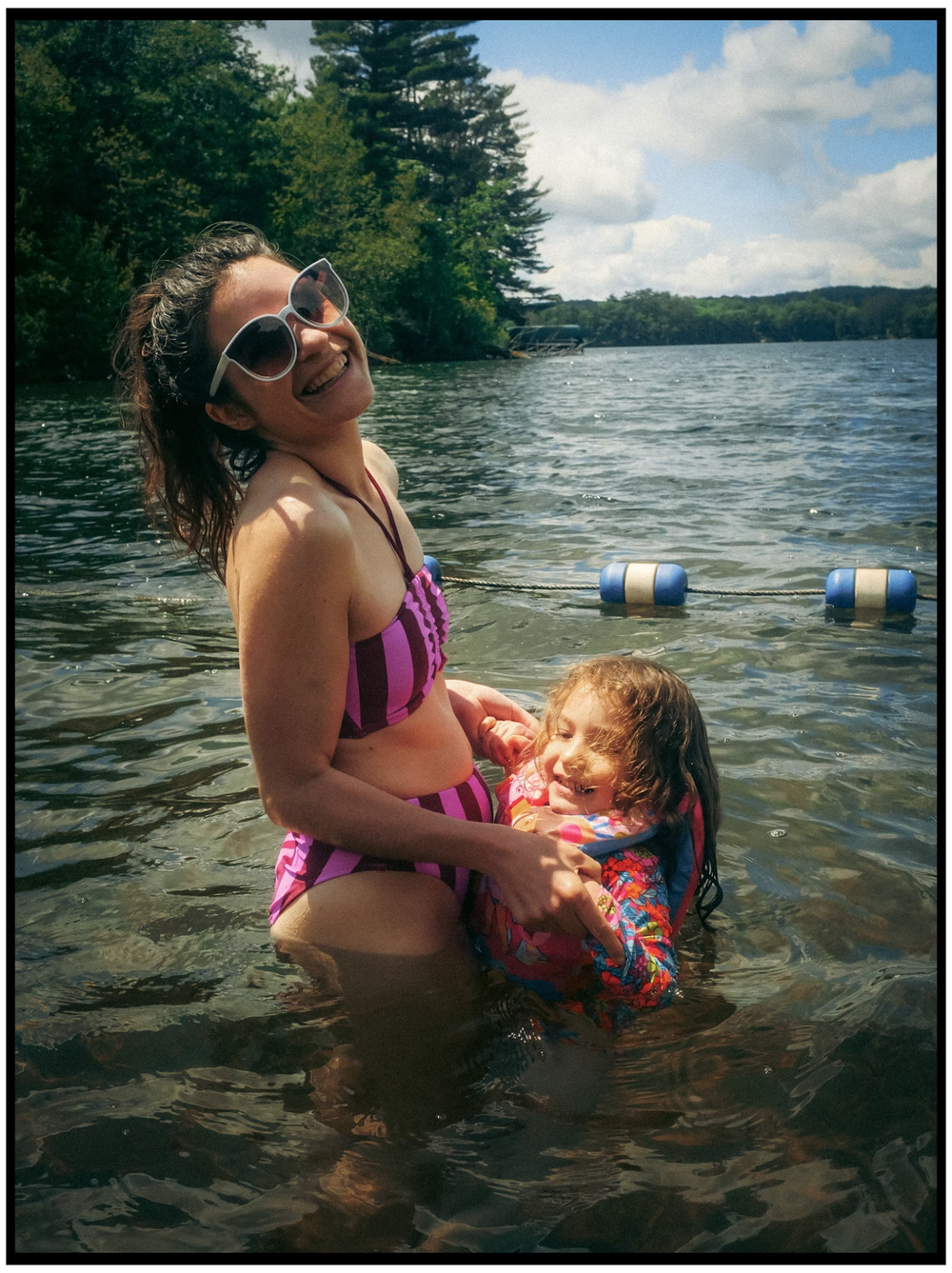 A sunny day at the lake shows people swimming and enjoying the water with trees and blue sky in the background.