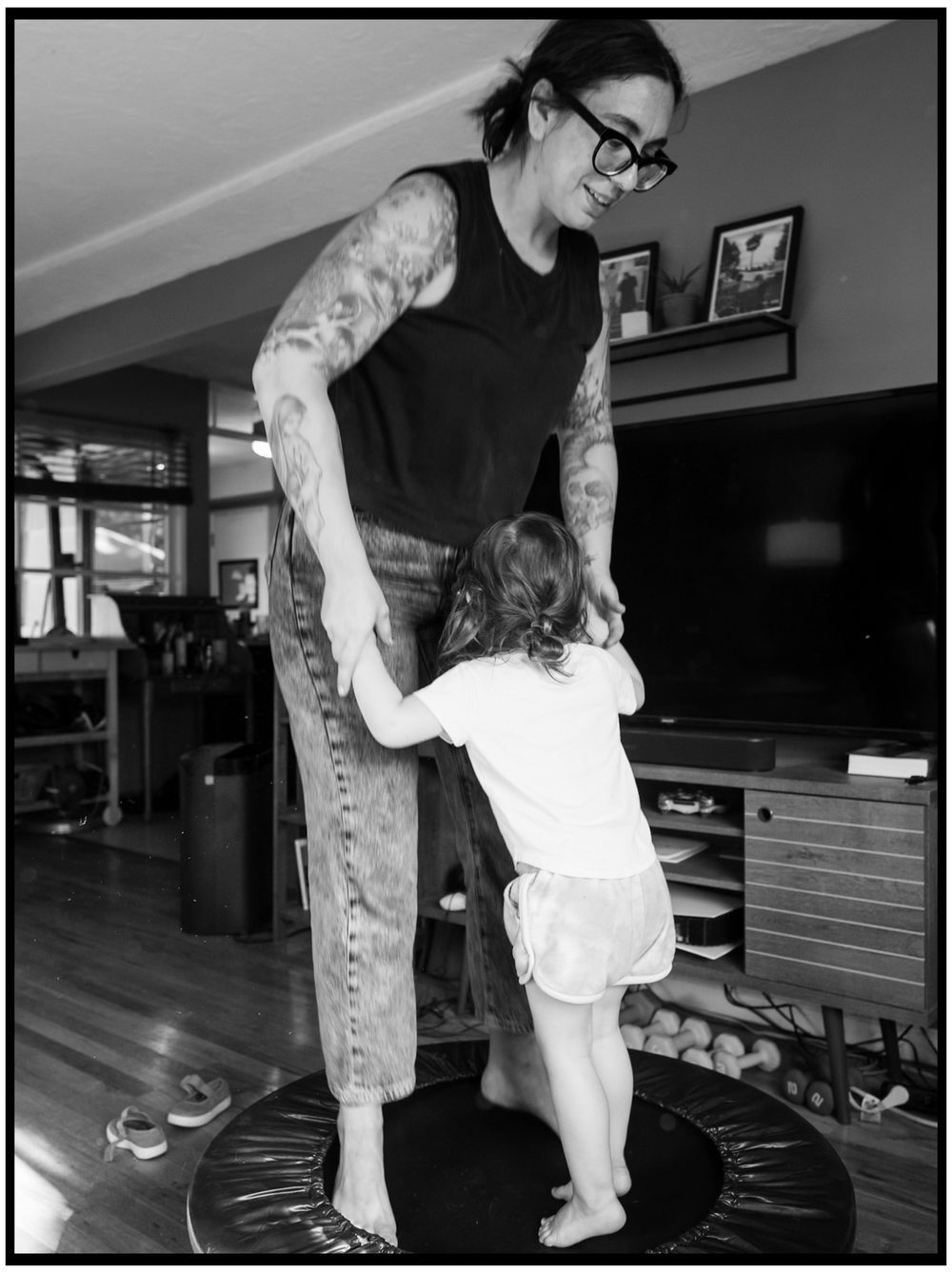A black and white photo of an adult and small child holding hands while standing on a mini trampoline indoors.