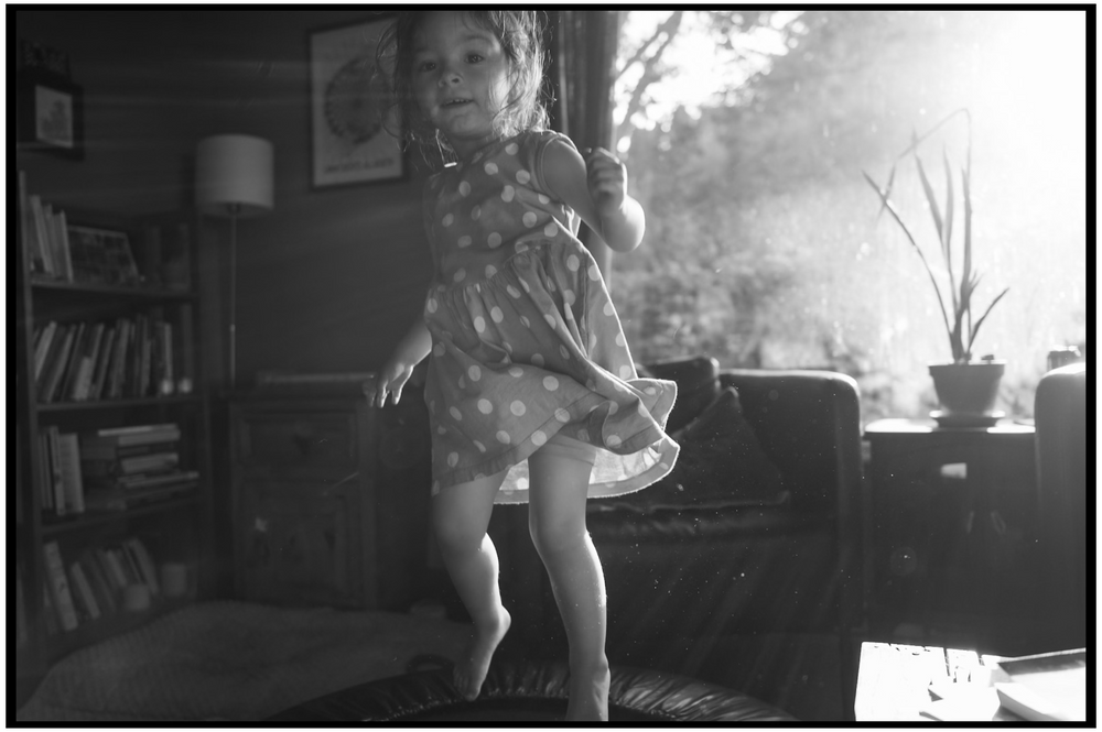 A young child dances in sunlit living room with bookshelves and houseplants visible in background.