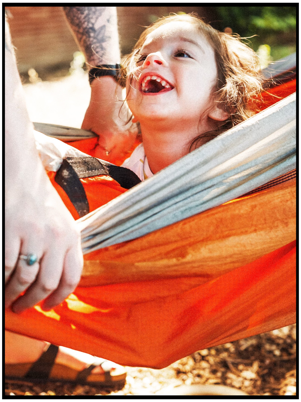 A child lying in an orange hammock laughs joyfully while relaxing outdoors on a sunny day.