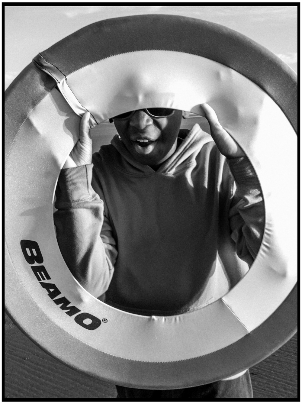 A black and white artistic photograph shows a subject looking through a white ring light with Seamo branding.