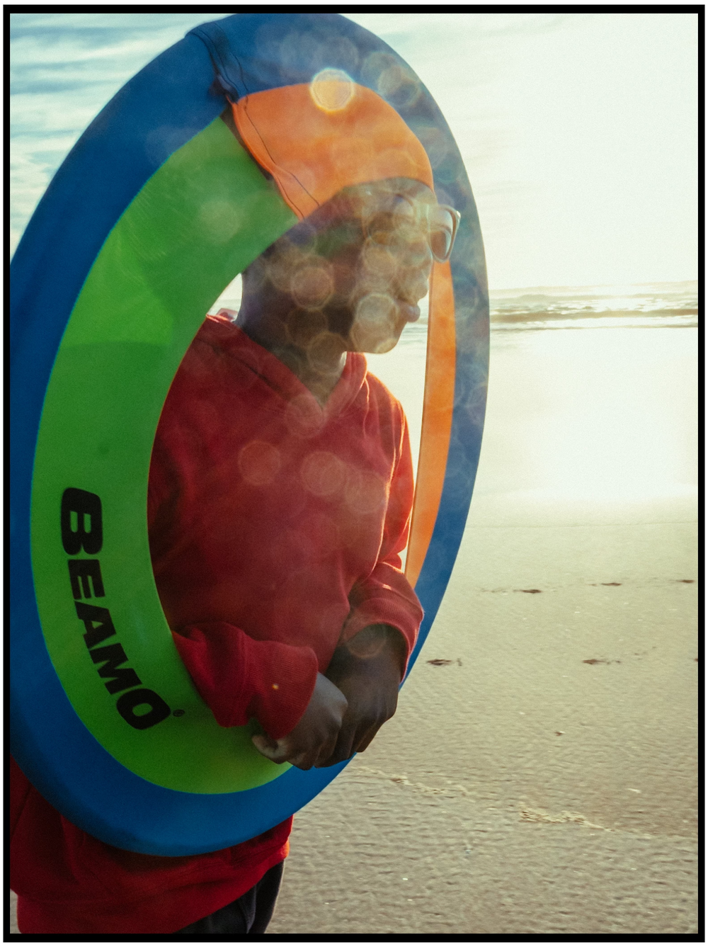 A person holds a colorful blue and green boogie board on a beach during sunset creating a silhouetted profile.