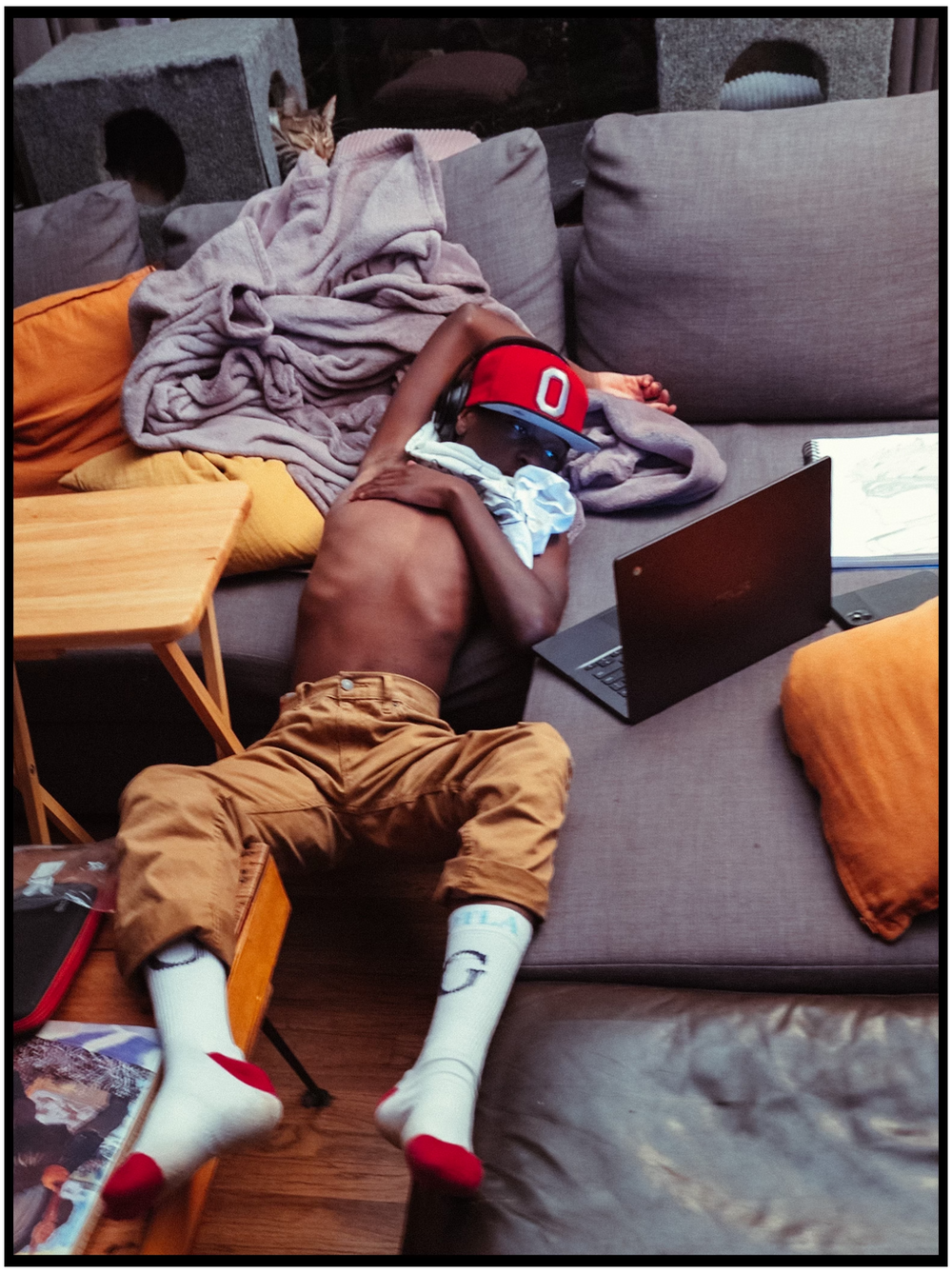 A tired person napping on a gray couch with scattered pillows and a baseball cap, wearing khaki shorts and socks.