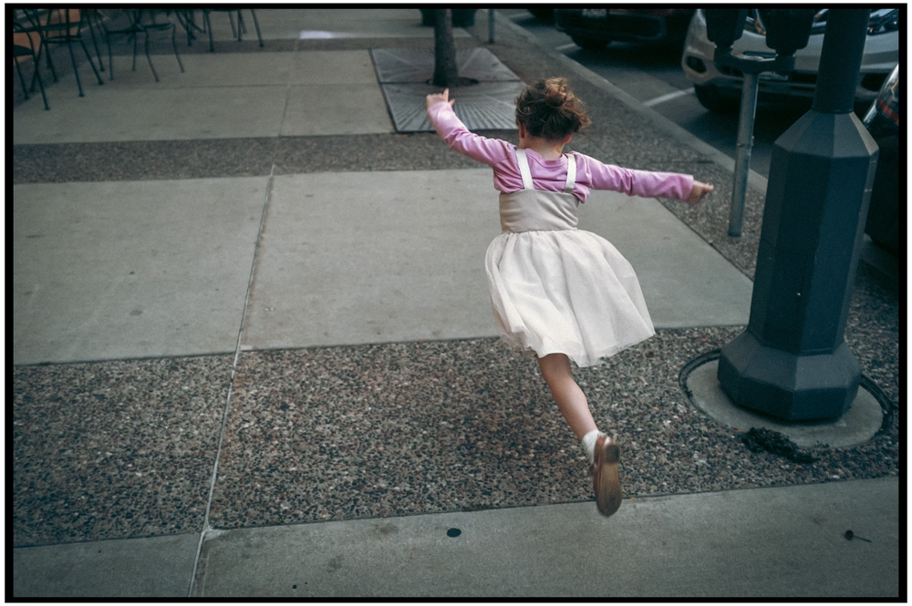 Person in pink shirt and white skirt dances gracefully on a city sidewalk with arms outstretched.