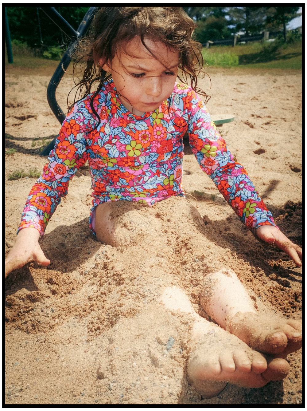 Child in colorful floral shirt plays in the sand at a playground on a sunny day, digging with bare feet visible.