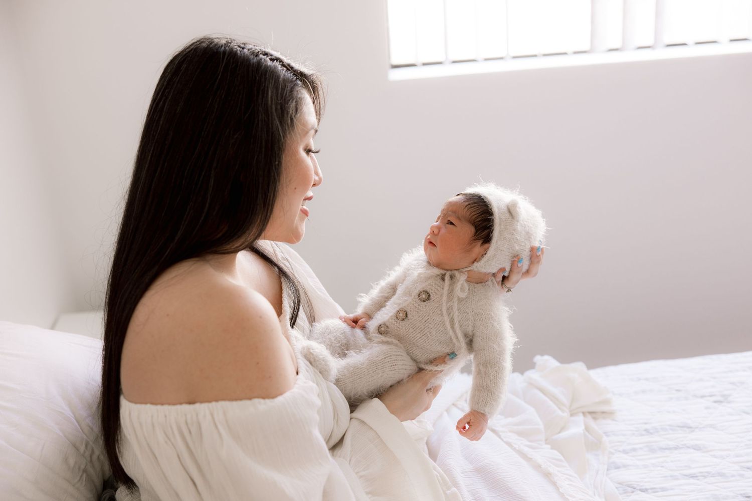 Parent and baby share a joyful moment together on a white bed in a sunlit room.