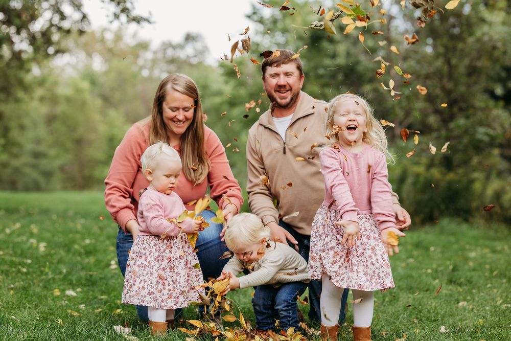 Family in coordinating pink and brown outfits plays with autumn leaves in a green park setting.