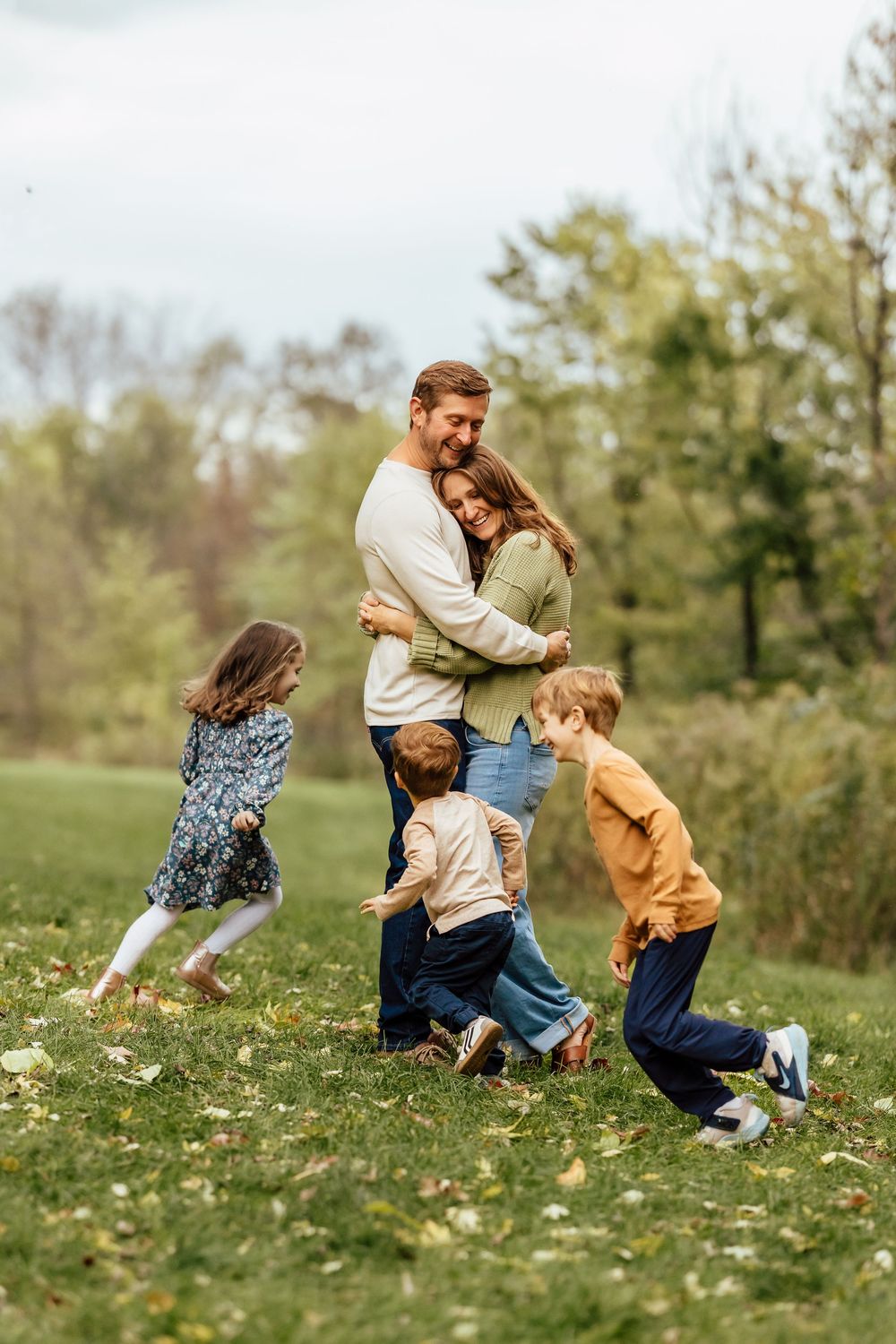 A family enjoys a playful moment together on a grassy hillside surrounded by trees on an autumn day.